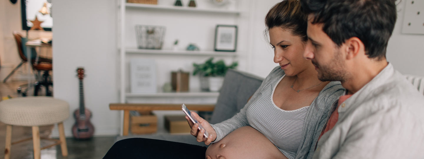 Eine Schwangere und ihr Mann sitzen auf dem Sofa und informieren sich mobil über eine Schwangerschaftsvorsorge.