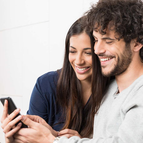 Ein junges Paar sitzt auf der Couch und schaut lächelnd auf ein Handy